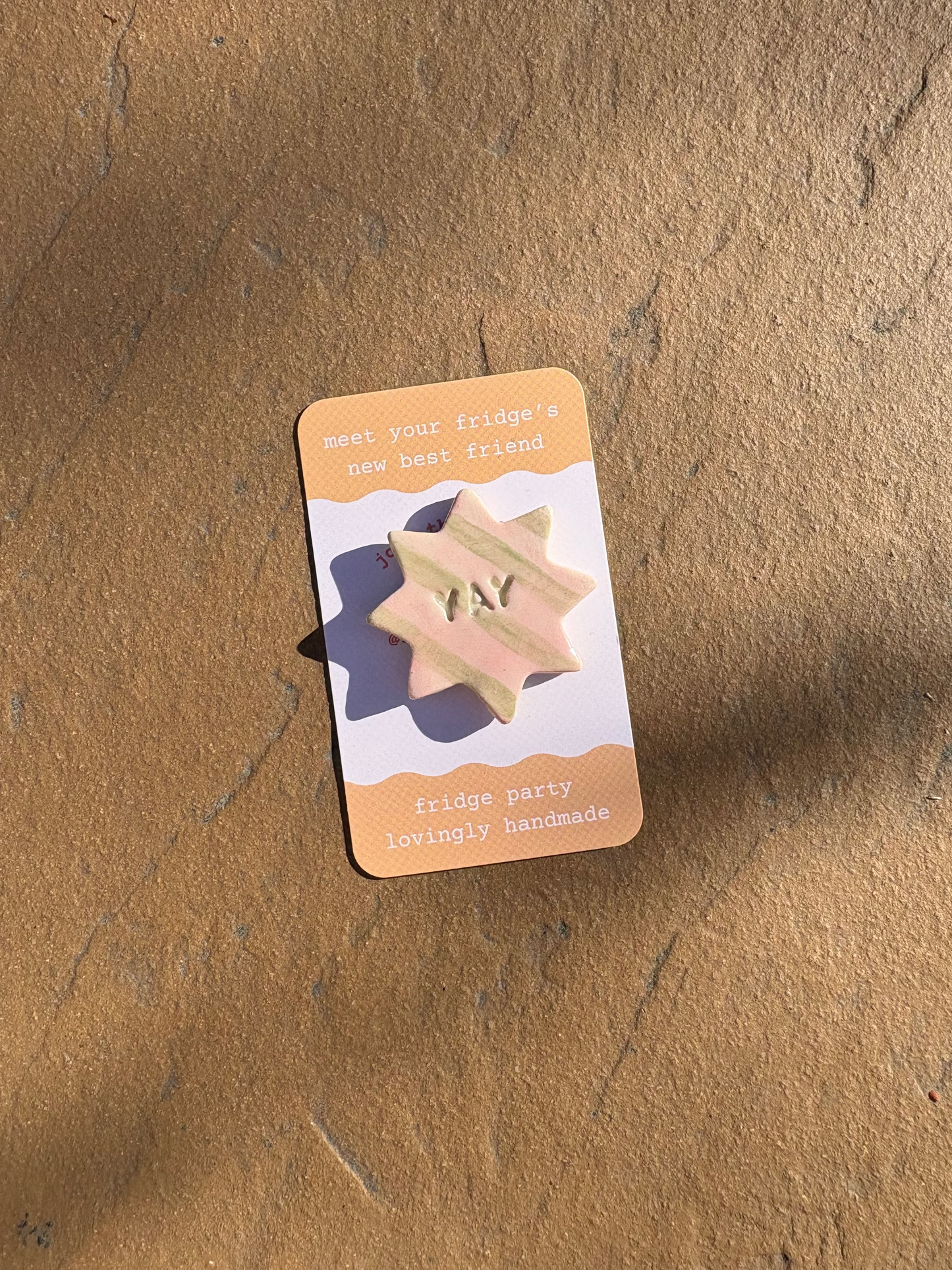 A small star-shaped pink and green striped fridge magnet with the word 'YAY' embossed in the centre, mounted on a white card with peach-colored borders. The card is placed on a textured brown surface with soft shadows.